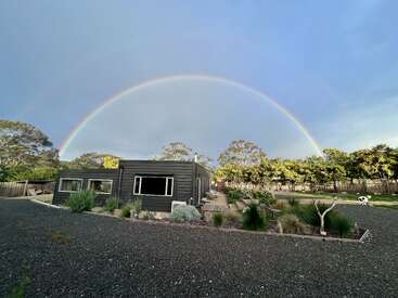 Ein modernes Haus steht in einem angelegten Garten unter einem lebendigen, bunten Regenbogen, der sich über den Himmel wölbt, umgeben von Bäumen und einem Schotterplatz. Eine friedliche Szene.