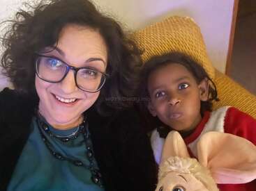 The image shows a woman and a child sitting together on a yellow couch, with the child holding a stuffed animal. The woman is wearing glasses.