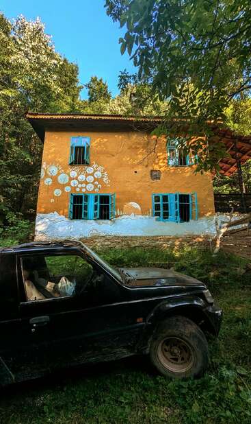 Una rústica casa amarilla con ventanas azules luce un caprichoso diseño artístico blanco en las paredes. Rodeada de exuberante vegetación, un viejo coche negro está aparcado delante. Una escena tranquila y serena.