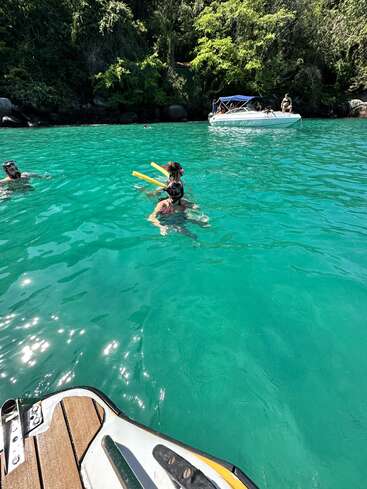Tres personas bucean en aguas turquesas cerca de una exuberante costa verde. Un barco blanco flota cerca, bajo la brillante luz del sol. La escena es tropical y relajada.