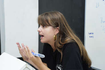 Una mujer de pelo largo y castaño, con pendientes de aro dorados y camisa negra, gesticula expresivamente mientras sostiene un rotulador, posiblemente enseñando o explicando algo apasionadamente.