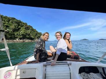 La imagen muestra a tres mujeres en un barco en el océano, con una isla verde al fondo y un cielo azul en lo alto.