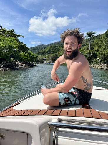 La imagen muestra a un hombre sentado en una barca en una serena masa de agua, rodeado de exuberante vegetación y un cielo azul con nubes blancas.