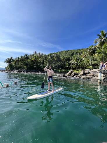 La imagen muestra a un hombre de pie sobre una tabla de paddleboard en el océano, con un remo en la mano, con palmeras y una costa rocosa de fondo en un día soleado.