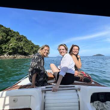 L'image représente trois femmes sur un bateau dans l'océan, avec une île verdoyante à l'arrière-plan et un ciel bleu au-dessus.
