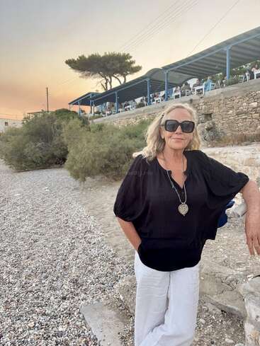 Eine Frau mit blonden Haaren und einer großen Sonnenbrille steht bei Sonnenuntergang auf einem Kieselstrand, ganz in Schwarz gekleidet, mit einem Restaurant und Bäumen im Hintergrund.