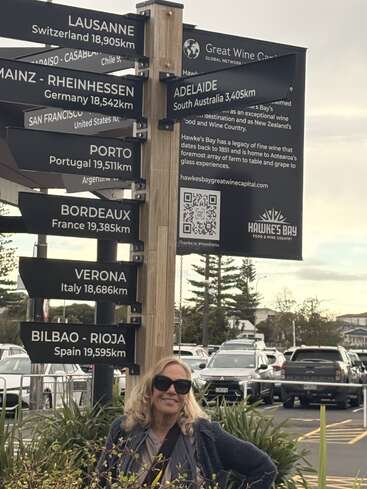 Dans un parking, une femme souriante portant des lunettes de soleil pose devant un panneau indiquant les distances vers des régions viticoles célèbres, notamment Porto, Bordeaux, Vérone et Bilbao-Rioja.