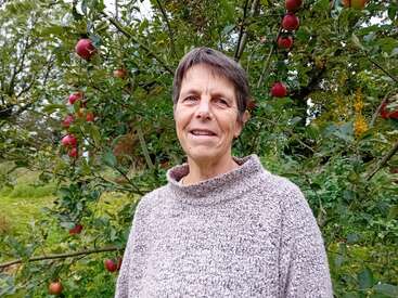 Una persona con el pelo corto y un jersey gris posa sonriente delante de un manzano, rodeada de follaje verde y manzanas rojas maduras al aire libre.