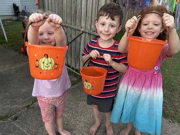 Three cheerful children hold orange Halloween buckets, smiling brightly. They're standing outdoors near a wooden fence, dressed casually, ready for trick-or-treat fun and candy collecting!