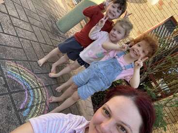Three smiling children and a woman pose for a cheerful selfie outside, making peace signs near a colorful chalk-drawn rainbow on a sunny paved area.