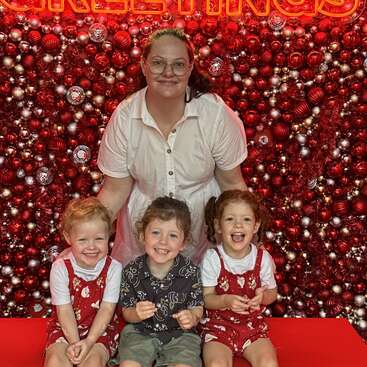 A woman in a white dress poses with three smiling children in front of a festive, red, and silver Christmas ornament backdrop with "GREETINGS" in neon letters.