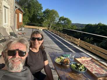 A smiling couple wearing sunglasses sits on a sunlit wooden deck, enjoying a meal of salad and pizza with beautiful green trees and hills in the background.