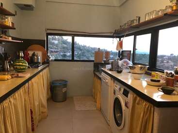 La imagen muestra una cocina compacta de paredes y encimeras blancas, con ventana, fregadero, fogones, lavadora y diversos utensilios y electrodomésticos.