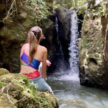 A woman sits on a mossy rock, gazing at a small waterfall cascading into a pool of water, surrounded by lush greenery and rocks.