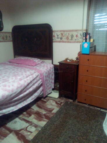 Um quarto aconchegante com uma cama de madeira, roupa de cama com estampa floral rosa, uma mesinha de cabeceira, uma cômoda, uma faixa decorativa no papel de parede, piso de mármore e uma janela com cortinas.