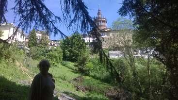 Uma pessoa está parada à sombra das árvores numa encosta verdejante, com um grande edifício histórico e uma torre visíveis ao fundo, rodeados por uma vegetação exuberante.