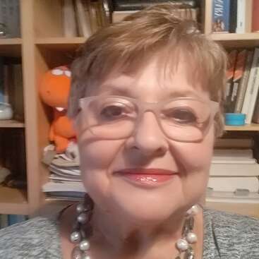 Uma senhora sorridente, de cabelo curto e óculos, está em frente a uma estante cheia de livros. Ela está vestindo uma blusa cinza e um colar de pérolas bem grande. Um bichinho de pelúcia laranja está atrás dela.