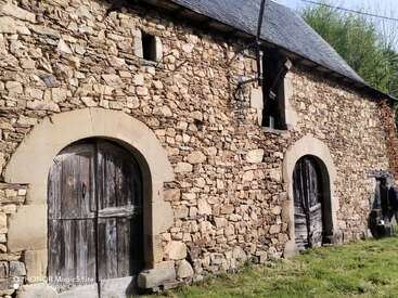 Cette image montre une vieille grange en pierre avec des portes en bois cintrées, du bois patiné et un toit en ardoise. On aperçoit de l'herbe et une personne près du bâtiment.