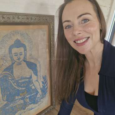 Una mujer sonriente con el pelo largo y castaño posa junto a una obra de arte azul enmarcada que representa una figura serena de Buda, que irradia energía pacífica. El decorado aparece en un interior.
