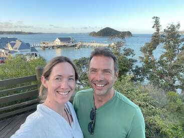 Das Bild zeigt ein Paar, das ein Selfie auf einer Terrasse mit Blick auf ein ruhiges Gewässer macht, mit einem malerischen Steg und Gebäuden im Hintergrund.