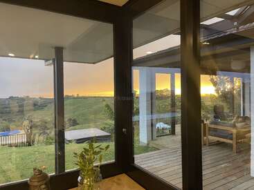 A imagem retrata uma cena serena do interior e exterior de uma casa, com uma grande janela com vista para um quintal, deck e pôr do sol.