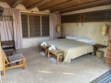 This image shows a rustic bedroom with a large bed, wooden benches, woven chairs, beige curtains, simple decor, natural light, and an earthy, minimalist ambiance.