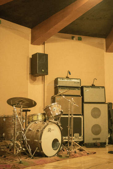 A cozy music studio with wooden beams, features a vintage Rogers drum set, stacked Fender amplifiers, large speakers, and a wall-mounted monitor speaker, all warmly lit.