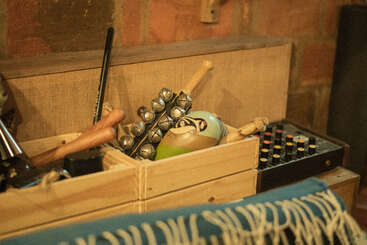 Wooden boxes contain various musical instruments, including maracas and jingle bells, beside a sound mixer. The scene is cozy with warm lighting and a patterned blanket.