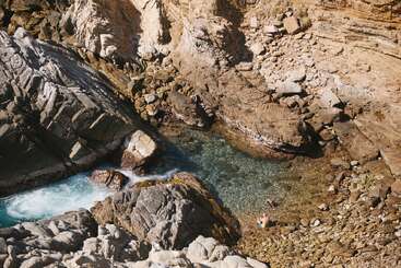 A rocky coastal area with large stone formations surrounds a small, clear tidal pool. Two people are relaxing in the shallow water, enjoying nature's tranquility.