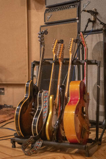 Five electric guitars rest on a stand in a recording studio, with vintage amplifiers and a microphone in the background, capturing a creative music atmosphere.