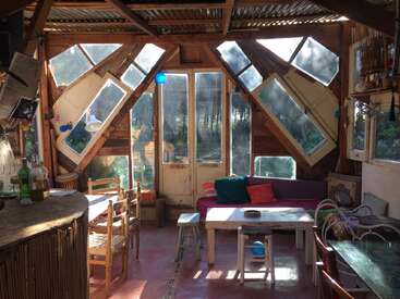 La imagen muestra una acogedora habitación con sofá, mesa, sillas y ventanas, con un interior rústico de madera y una serena vista al exterior a través de los grandes ventanales.