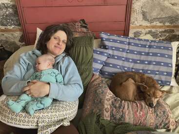 Une femme se détend sur un lit, un bébé blotti dans ses bras, tandis qu'un petit chien brun dort paisiblement à côté d'elle, sur un tas d'oreillers. Une scène chaleureuse.