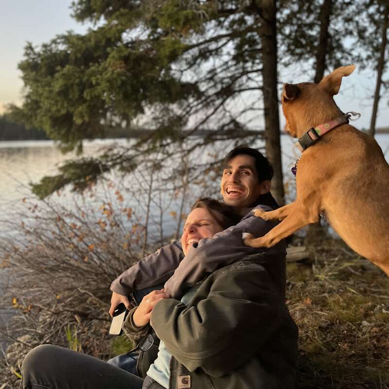 La imagen representa una escena serena de un hombre y una mujer sentados junto a un lago, con un perro sobre los hombros del hombre, todos vestidos con ropa informal.