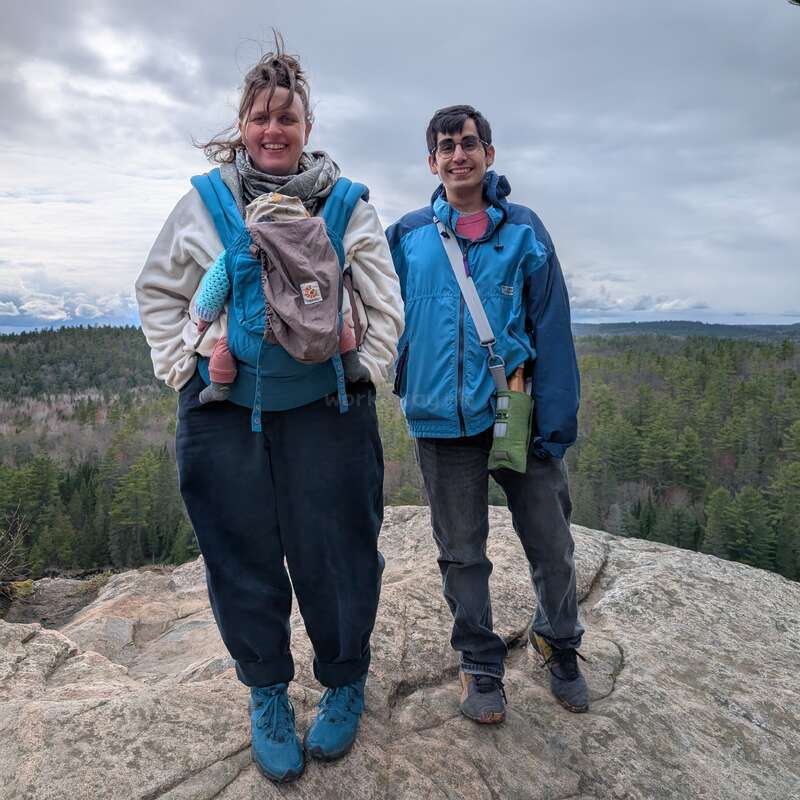 Dois adultos, um deles carregando um bebê em um carrinho de bebê frontal, estão no topo de um penhasco rochoso com vista para uma vasta paisagem florestal, sob um céu nublado. Rostos sorridentes.