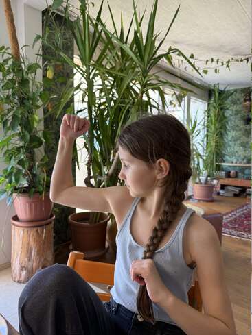 Una chica con una larga trenza flexiona el brazo para demostrar su fuerza. Está sentada en el interior, rodeada de exuberantes plantas verdes, y lleva una camiseta sin mangas azul.