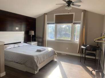 Esse quarto tem uma cama confortável com roupa de cama listrada, janela grande com luz solar, parede escura de destaque, criado-mudo, abajur, escrivaninha com cadeira e cabideiro.