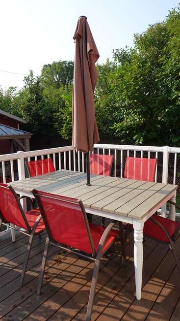 A patio scene shows a wooden table with six red chairs, a closed brown umbrella, wooden deck flooring, white railing, and lush green trees surrounding the area.