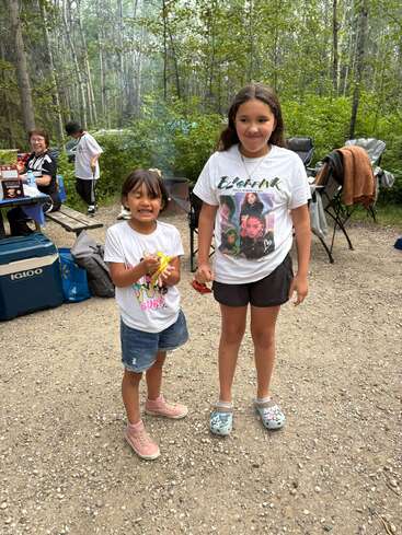 Two smiling girls stand on a gravel path at a forest campsite. Behind, people sit by a picnic table, surrounded by camping gear and tall trees.