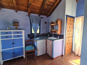 Essa cozinha rústica aconchegante tem armários azuis, teto de madeira, uma pequena pia, geladeira, janela com cortinas claras, azulejos de terracota e uma iluminação natural aconchegante e convidativa.