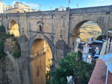 A imagem mostra uma impressionante ponte de pedra que atravessa um desfiladeiro profundo, com arcos e vista para um terraço panorâmico abaixo. A luz do sol ilumina a estrutura antiga.