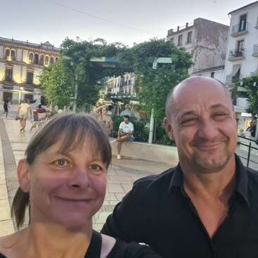 Um casal sorridente tira uma selfie em uma animada praça pública ao anoitecer. As pessoas relaxam em bancos e passeiam, cercadas por vegetação e charmosos edifícios antigos.