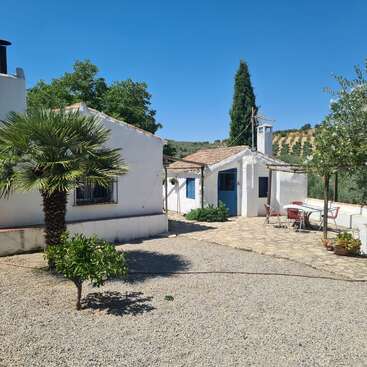 Uma charmosa casa de campo com paredes brancas, portas azuis e assentos ao ar livre. Cercada por árvores, plantas e um pátio de cascalho sob um céu azul brilhante.