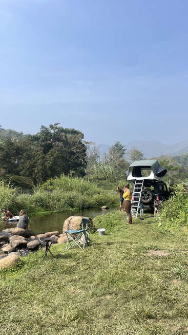 Un grupo de personas disfruta de una acampada junto a un río tranquilo, con una tienda de campaña en el techo de un vehículo, rodeados de vegetación y montañas bajo un cielo azul y despejado.