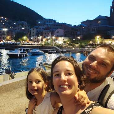The image depicts a family of three posing for a selfie in front of a picturesque harbor at dusk, with boats and buildings in the background.