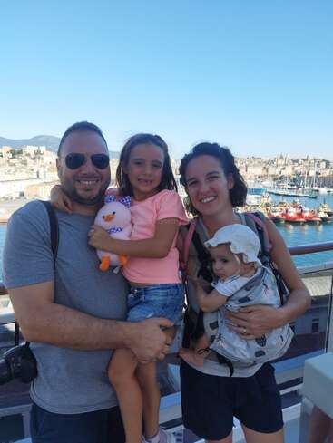 A smiling family of four poses outdoors on a sunny day, with a scenic harbor and city in the background. The children look happy and cheerful.