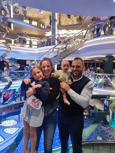 A happy family of four poses, smiling, inside a luxurious, modern, multi-level building with glass, spiral railings, and reflections, surrounded by people in the background.
