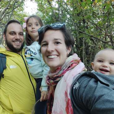 A happy family of four poses for a selfie outdoors, surrounded by trees. Everyone is smiling, except the girl, who makes a surprised facial expression.
