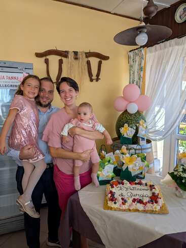 A happy family of four poses together indoors. The parents smile warmly, holding their two daughters, one older girl in pink sequins, the baby wearing pink.