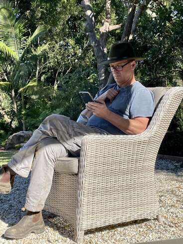 The image depicts a man sitting in a wicker chair, engrossed in his phone, with a serene garden backdrop featuring trees and lush greenery.
