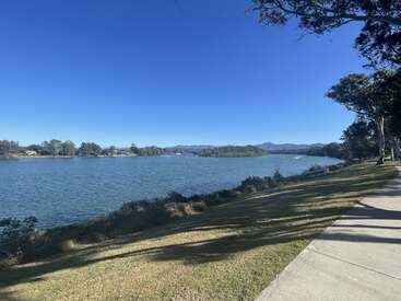 The image depicts a serene lake scene with a concrete path, lush grass, and trees, set against a clear blue sky and distant mountains on a sunny day.